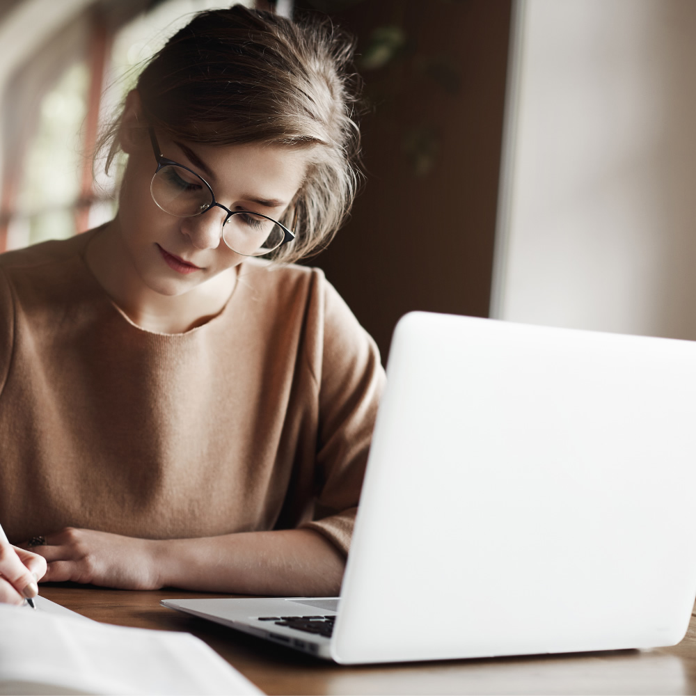 Imagen de portada de curso "Inmersión en habilidades y competencias digitales" - Se visualiza una Mujer con anteojos, observando una pantalla de notebook y anotando con birome en un papel