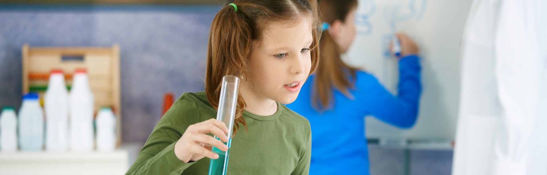 Imagen de portada de curso "Pensamiento científico en el Nivel Primario" - Se visualiza una preadolescente con un tubo de ensayo en la mano y otra chica escribiendo en un pizarrón