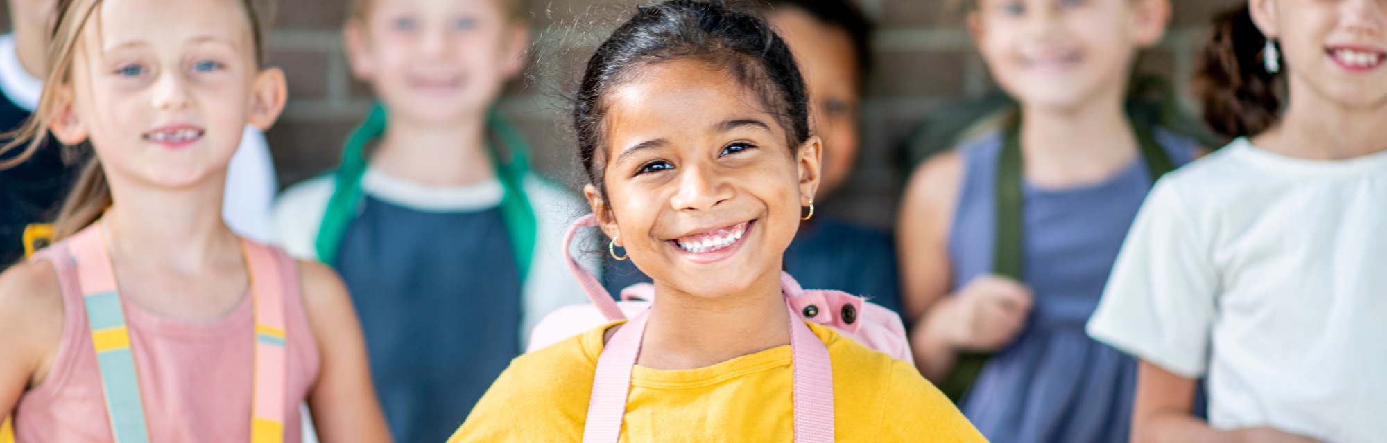 Imagen de portada del curso "Variables que influyen en el aprendizaje: ¿cómo lograr experiencias significativas en los alumnos rediseñando los espacios, tiempos y recursos?" - Se visualiza una Chica sonriendo, mirando hacia el frente y sus compañeros en el fondo, sonriendo también.